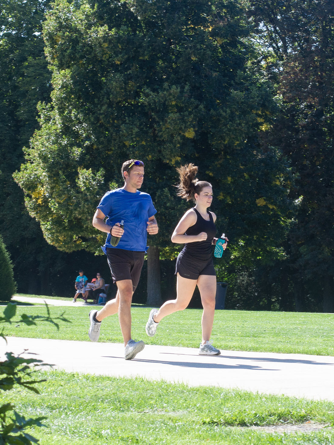 Un homme à t-shirt bleu et short noir coure avec une femme en habits de sports noirs, tout les deux avec une gourde running à la main
