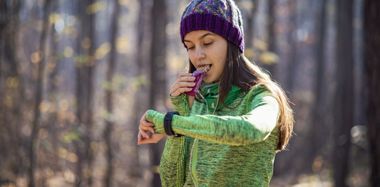 une femme sportive est dans une foret, elle est en arrêt car elle mange une barre proteinees, elle a un gilet vert, un bonnet violet et une montre connecté