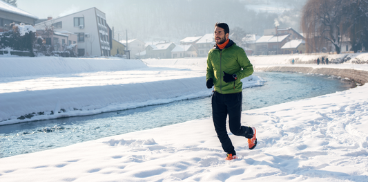 homme en train de courir près d'un ruisseau dans la nature près d'une forêt, il est habillé avec une veste verte et un jogging noir, avec des baskets de running noires et oranges